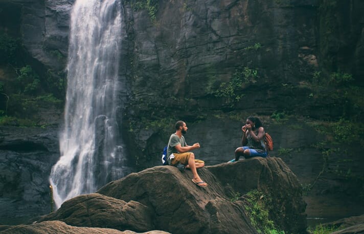 An image of two people sitting on rocks in front of a waterfall and rock wall.